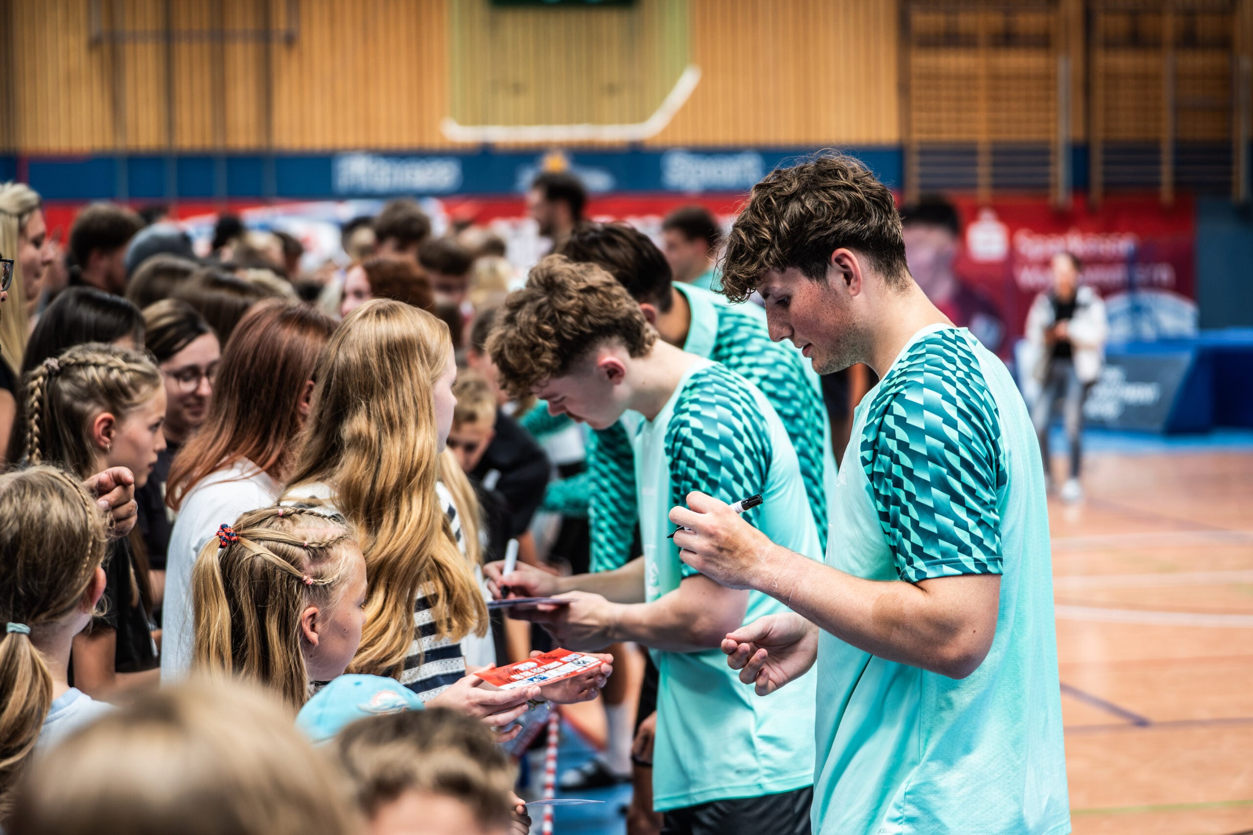 Mehrere Sportler in blaugrünen Trikots geben einer Schar junger Fans in einer Sporthalle Autogramme. Es herrscht reges Treiben, und die Fans halten eifrig Gegenstände zum Signieren hin – eine lebhafte Atmosphäre, die an den Moment erinnert, als Neu-Trainer Börge Lund beim THW Kiel vorgestellt wurde.