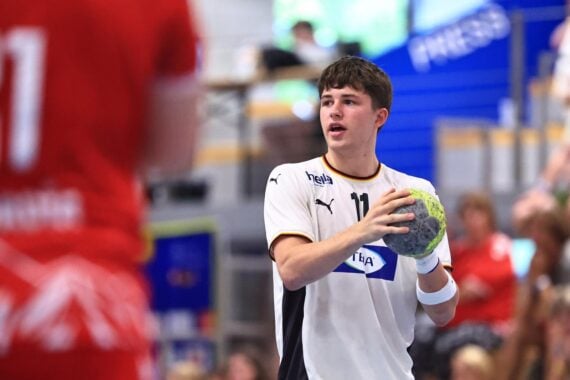 Ein junger Handballspieler in einem weißen Trikot, der für den THW Kiel antritt, hält einen Ball in der Hand und bereitet sich während eines Spiels der Handball-Bundesliga-Saison 2026/2027 auf einen Wurf vor. Im Vordergrund ist ein weiterer Spieler in Rot unscharf zu sehen, während im Hintergrund Zuschauer und der kürzlich ernannte Trainer Börge Lund zu erkennen sind.