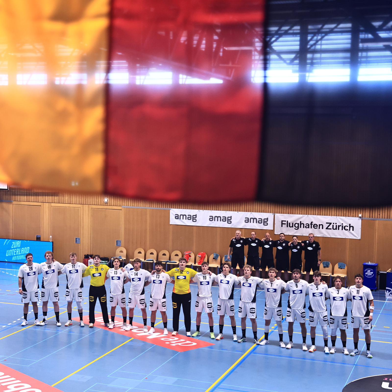 Eine Handballmannschaft in weißen Trikots steht in einer Reihe auf einem Hallenfeld, die Arme umeinander gelegt, während darüber teilweise eine große schwarz-rot-goldene Fahne zu sehen ist. Im Hintergrund sind Trainer – darunter der neue Trainer des THW Kiel für die Handball-Bundesliga-Saison 2026/2027, Börge Lund – sowie Transparente zu sehen.