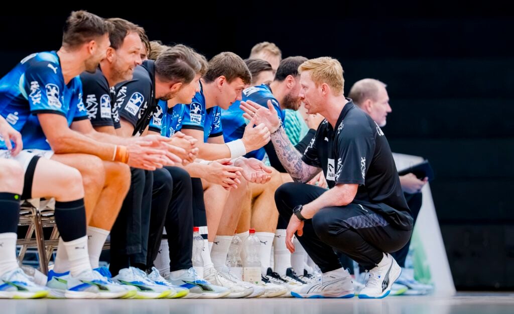 HANDBALL 2026/27: THW Kiel im Trainingslager in Graz Ein Trainer hockt vor sitzenden männlichen Sportlern in blauen Trikots und gibt ihnen während eines Spiels Anweisungen. Die Spieler hören aufmerksam zu; in ihrer Nähe sind Wasserflaschen und Ausrüstung zu sehen.