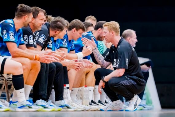 Ein Trainer hockt vor sitzenden männlichen Sportlern in blauen Trikots und gibt ihnen während eines Spiels Anweisungen. Die Spieler hören aufmerksam zu; in ihrer Nähe sind Wasserflaschen und Ausrüstung zu sehen.