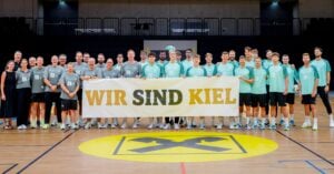 Eine große Gruppe von Menschen in Sportkleidung steht in einer Sporthalle und hält ein Transparent mit der Aufschrift „WIR SIND KIEL“ hoch. Auf dem Boden ist ein großes gelbes Logo zu sehen, und im Hintergrund sind leere Tribünen zu erkennen.