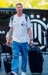 Ein Mann in einem weißen T-Shirt und blauen Jeans lächelt, während er zwei Koffer hinter sich herzieht. Er trägt einen Rucksack, hat sichtbare Tattoos und steht vor einem schwarzen Bus mit weißer Beschriftung und Sternen.