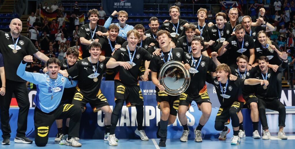 F - Germany vs Slovenia A team of athletes and coaches celebrate their victory by posing with a trophy on an indoor sports court, surrounded by cheering spectators and national flags.
