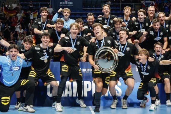 A team of athletes and coaches celebrate their victory by posing with a trophy on an indoor sports court, surrounded by cheering spectators and national flags.