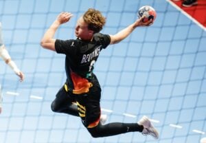An athlete leaps mid-air to pass a ball during an indoor handball game, while teammates and opponents are positioned on the court.