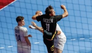 A player in a black jersey with the number 11 leaps to strike a ball during a handball match, while an opponent in a white jersey watches closely.