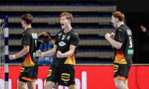 A group of handball players in black and orange uniforms celebrate a goal on the court, while an opposing player in white stands nearby.