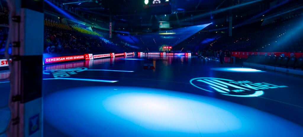 HANDBALL 2025/26: Aktion „Euer Topspiel“ Eine hell beleuchtete Sporthalle mit leeren Sitzplätzen, blauen Scheinwerfern auf dem Spielfeld, digitalen Werbeanzeigen an den Seiten und einer im Hintergrund sichtbaren Anzeigetafel.