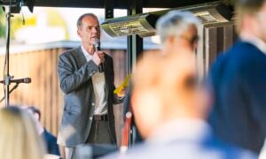 Ein Mann in einem grauen Anzug spricht bei einer Veranstaltung im Freien in ein Mikrofon und hält dabei ein gelbes Blatt Papier in der Hand. Andere Personen im Vordergrund und Hintergrund sind unscharf.