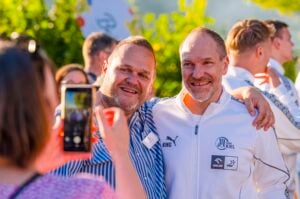 Zwei lächelnde Männer posieren bei einer Veranstaltung im Freien für ein Foto; der eine trägt ein gestreiftes Hemd, der andere eine weiße Sportjacke. Eine Person im Vordergrund fotografiert sie mit einem Smartphone. Im Hintergrund sind weitere Personen zu sehen.