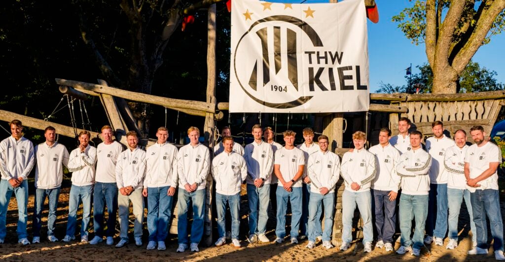Eine Gruppe von Männern in weißen Jacken und blauen Jeans steht draußen vor einem großen THW-Kiel-Banner und posiert im Sonnenlicht für ein Mannschaftsfoto.