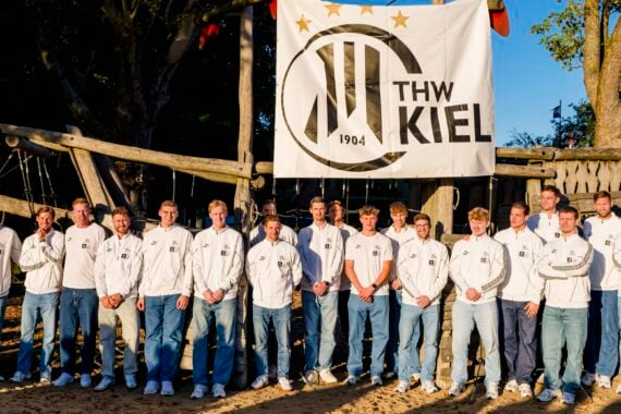 Eine Gruppe von Männern in weißen Jacken und blauen Jeans steht draußen vor einem großen THW-Kiel-Banner und posiert im Sonnenlicht für ein Mannschaftsfoto.