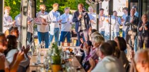 Ein Mann spricht in ein Mikrofon, während hinter ihm eine Gruppe junger Menschen in weißen Hemden steht und klatscht. Die an langen Tischen sitzenden Gäste beobachten das Geschehen und applaudieren in einer sonnendurchfluteten, festlichen Atmosphäre.