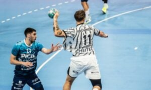 Zwei männliche Handballspieler treten auf einem blauen Hallenfeld gegeneinander an: Der eine in blauem Trikot bereitet sich auf die Verteidigung vor, während der andere, in Weiß und Schwarz, den Ball hoch in die Luft hält und bereit ist, einen Spielzug zu starten.