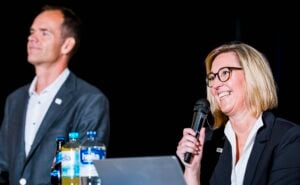 Eine Frau im Anzug lächelt, während sie neben einem Laptop und Wasserflaschen in ein Mikrofon spricht. Im Hintergrund steht ein Mann im Anzug, der jedoch unscharf zu sehen ist. Beide befinden sich bei einer Veranstaltung in einem Raum mit dunklem Hintergrund.