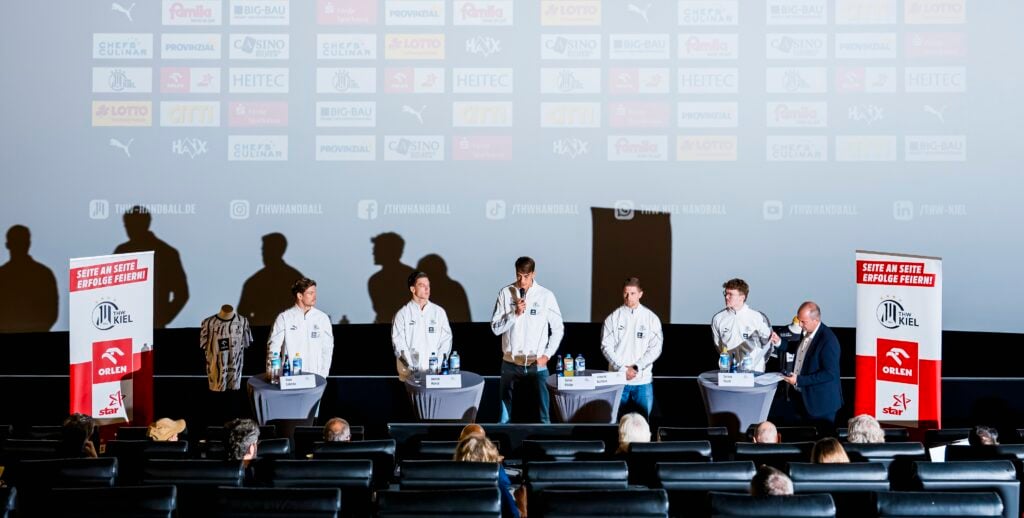 Fünf Personen stehen mit Mikrofonen an einem Podiumstisch vor einem Kinopublikum; hinter ihnen werden auf einer Leinwand Firmenlogos gezeigt, und zu beiden Seiten sind jeweils zwei rote Banner aufgestellt.