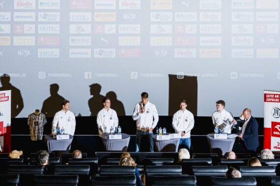 Fünf Personen stehen mit Mikrofonen an einem Podiumstisch vor einem Kinopublikum; hinter ihnen werden auf einer Leinwand Firmenlogos gezeigt, und zu beiden Seiten sind jeweils zwei rote Banner aufgestellt.