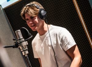 Eine Person mit Kopfhörern und einem weißen T-Shirt steht vor einem Mikrofon in einem Tonstudio, dessen Wände mit schalldämmenden Paneelen verkleidet sind.