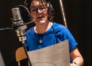 Eine Person mit Kopfhörern und einem blauen Hemd liest in einem Tonstudio vor einem Mikrofon von einem Skript ab.