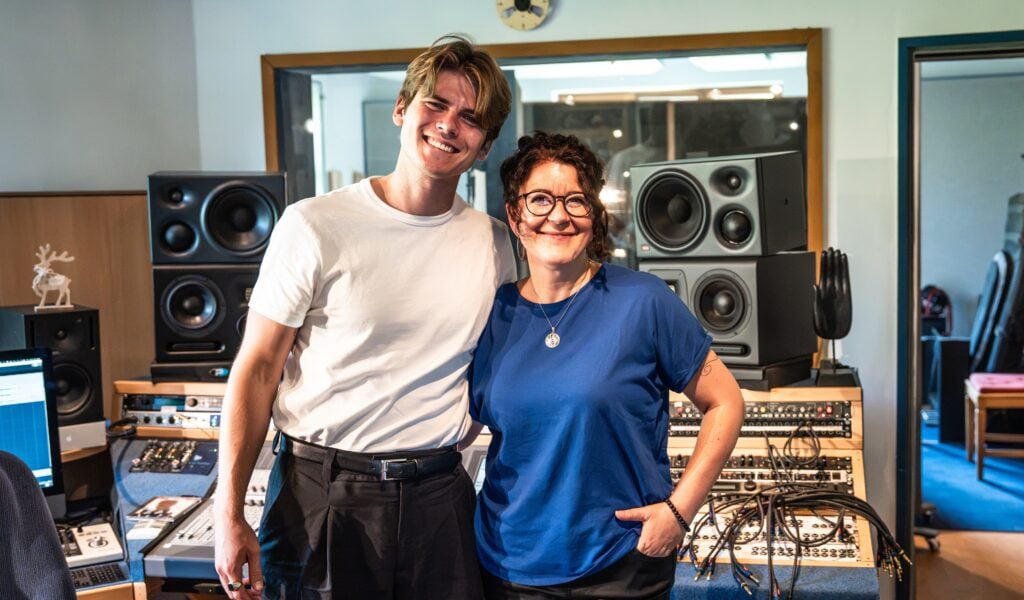 26_Tonstuido_Titel_2 Ein lächelnder Mann und eine lächelnde Frau stehen zusammen in einem Musikstudio, das mit Lautsprechern, Audiogeräten und Kabeln vollgestopft ist. Der Mann trägt ein weißes Hemd, die Frau ein blaues Hemd.