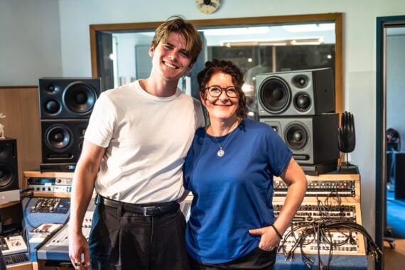 Ein lächelnder Mann und eine lächelnde Frau stehen zusammen in einem Musikstudio, das mit Lautsprechern, Audiogeräten und Kabeln vollgestopft ist. Der Mann trägt ein weißes Hemd, die Frau ein blaues Hemd.