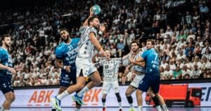 Ein Handballspieler in weißer Spielkleidung springt hoch, um den Ball zu werfen, während er von Verteidigern in blauer Spielkleidung umringt ist; im Hintergrund beobachtet eine Zuschauermenge das Geschehen.