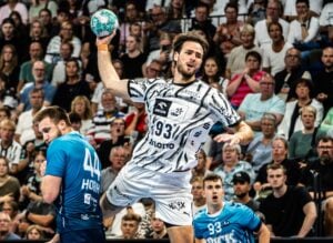 Ein Handballspieler in einem schwarz-weißen Trikot befindet sich in der Luft und bereitet einen Wurf vor, während zwei Spieler in blauen Trikots verteidigen. Eine große Zuschauermenge verfolgt das Geschehen gebannt.