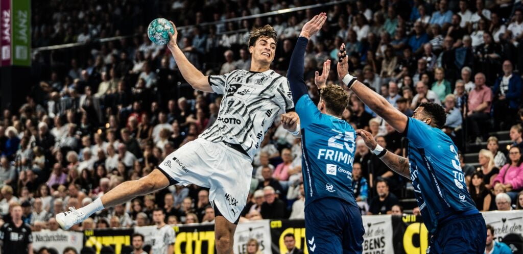 26_nachPick_koester Ein Handballspieler in weißem Trikot springt hoch, um den Ball zu werfen, während zwei Verteidiger in Blau versuchen, ihn zu blockieren – vor einer vollbesetzten Arena.