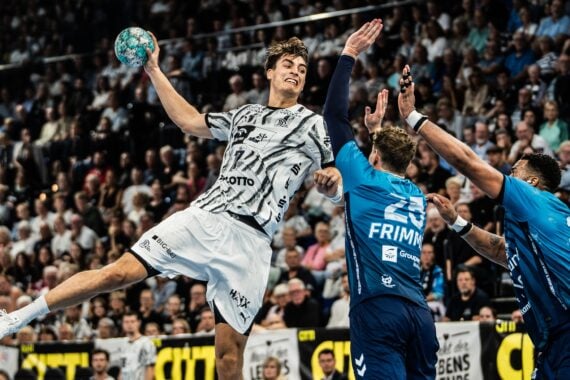 Ein Handballspieler in weißem Trikot springt hoch, um den Ball zu werfen, während zwei Verteidiger in Blau versuchen, ihn zu blockieren – vor einer vollbesetzten Arena.