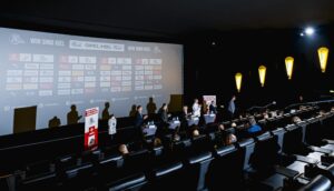 Eine Diskussionsrunde sitzt an einem Tisch auf der Bühne und blickt auf das Publikum in einem schwach beleuchteten Theater, während hinter ihr auf einer großen Leinwand verschiedene Sponsorenlogos zu sehen sind.