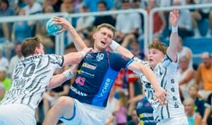 Drei männliche Handballspieler in Aktion; einer in Blau versucht zu werfen, während zwei in Weiß mit erhobenen Armen verteidigen. Im Hintergrund ist eine Zuschauermenge zu sehen, die das spannende Spiel verfolgt – genauso spannend wie damals, als der THW Kiel den Alpla-Sommercup in Hard gewonnen hat.