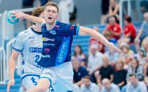 Ein Handballspieler im blauen Trikot springt hoch, um den Ball zu werfen, während ein Verteidiger im weißen Trikot versucht, ihn daran zu hindern. Im Hintergrund verfolgen Zuschauer das Geschehen von den Tribünen aus.