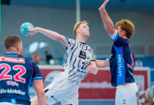 Ein Handballspieler in einem weißen Trikot springt hoch, um den Ball zu werfen, während er während eines Spiels von zwei Spielern in blauen Trikots verteidigt wird.