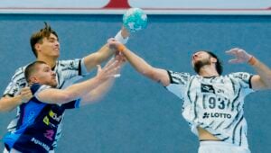 Drei männliche Handballspieler strecken in der Luft nach dem Ball; zwei in weißen Trikots und einer in Blau – alle sind während eines Hallenspiels hochkonzentriert.