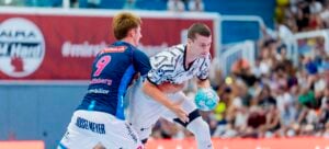 Zwei männliche Handballspieler kämpfen auf einem Hallenfeld um den Ball. Ein Spieler in einem schwarz-weißen Trikot hält den Ball, während der andere in Blau versucht, ihn zu blocken. Im Hintergrund verfolgen Zuschauer das Geschehen von den Tribünen aus.