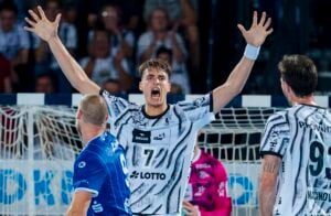 Ein männlicher Handballspieler in einem schwarz-weißen Trikot reckt während eines Spiels die Arme in die Höhe und jubelt, während sich in seiner Nähe Teamkollegen und Gegner befinden. Im Hintergrund sind die Zuschauer und das Tornetz zu sehen.