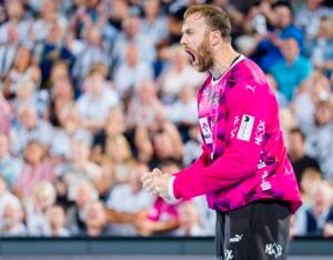 Ein Handballspieler in einem knallrosa Trikot jubelt während eines Spiels leidenschaftlich, ballt die Fäuste und schreit, während im Hintergrund eine verschwommene Menschenmenge zu sehen ist.
