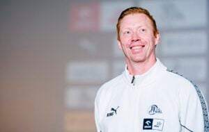 Ein lächelnder Mann mit heller Haut und roten Haaren trägt eine weiße Sportjacke mit verschiedenen Logos. Im unscharfen Hintergrund ist eine Wand mit undeutlichen Logos zu sehen.