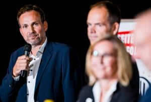 Ein Mann im Anzug hält ein Mikrofon in der Hand und hält bei einer Veranstaltung eine Rede. Zwei weitere Personen, ein Mann und eine Frau, stehen unscharf im Vordergrund. Im Hintergrund ist ein Banner teilweise zu sehen.