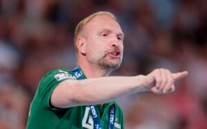 Ein Mann in einem grünen Hemd gestikuliert und zeigt beim Sprechen mit der rechten Hand; er wirkt dabei intensiv und konzentriert. Er trägt ein Schlüsselband, und der Hintergrund ist unscharf, was darauf hindeutet, dass er sich bei einer öffentlichen Veranstaltung oder in einer Sportstätte befindet.