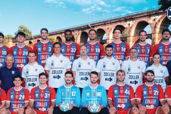 Eine Herren-Handballmannschaft in roten, blauen und weißen Trikots posiert im Freien vor einer großen Steinbrücke. Trainer und Betreuer stehen neben ihnen. In den oberen Ecken sind die Mannschaftslogos zu sehen.