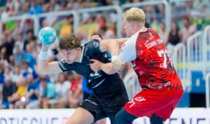 Zwei männliche Handballspieler liefern sich ein intensives Duell; der Spieler in Schwarz versucht, sich mit dem Ball in der Hand freizuspielen, während ein Spieler in Rot und Weiß ihn verteidigt. Im Hintergrund sind verschwommene Zuschauer zu sehen.
