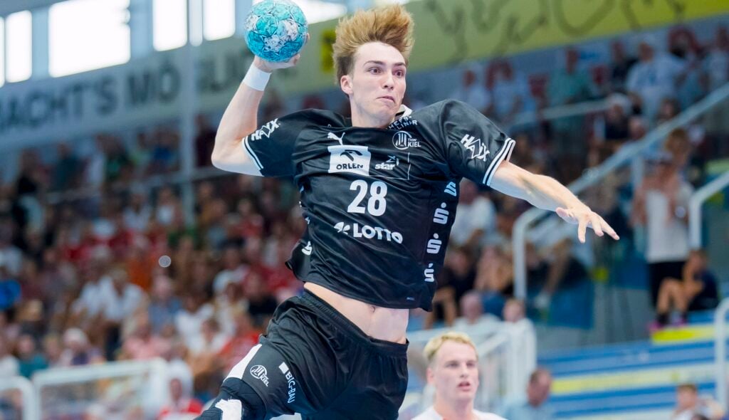 HANDBALL 2026/27: THW Kiel beim Alpla Sommercup Ein Handballspieler in schwarzem Trikot springt in die Luft, um den Ball zu werfen. Zuschauer verfolgen das Geschehen von den Tribünen aus, und hinter ihm ist ein weiterer Spieler in weißem Trikot zu sehen.