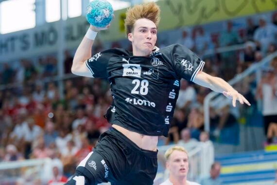 Ein Handballspieler in schwarzem Trikot springt in die Luft, um den Ball zu werfen. Zuschauer verfolgen das Geschehen von den Tribünen aus, und hinter ihm ist ein weiterer Spieler in weißem Trikot zu sehen.