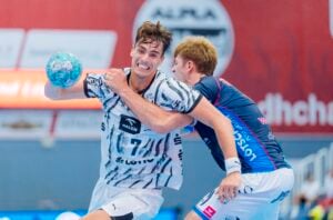 Zwei männliche Handballspieler liefern sich ein intensives Duell. Ein Spieler im weißen Trikot versucht, einen Wurf anzusetzen, während ein Gegner im blauen Trikot ihn am Arm packt, um ihn zu blockieren. Beide zeigen entschlossene Gesichtsausdrücke, die an die hart umkämpften Spiele erinnern, in denen der THW Kiel den Alpla-Sommercup in Hard gewonnen hat.