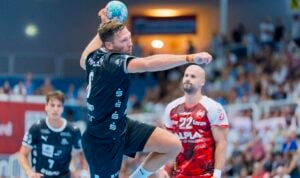 Ein Handballspieler in schwarzer Spielkleidung springt hoch, um den Ball zu werfen, während zwei gegnerische Spieler in roten und weißen Trikots während eines Spiels in einer vollbesetzten Halle verteidigen.