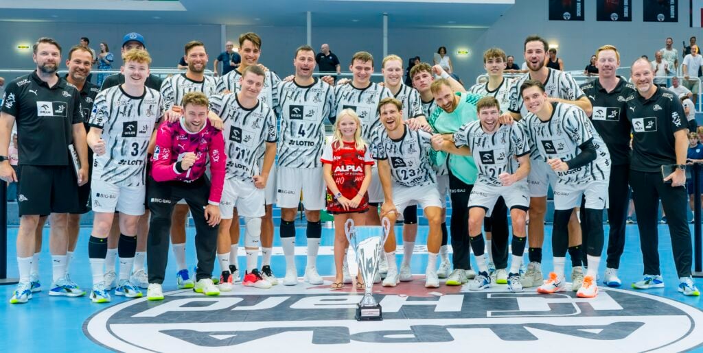 HANDBALL 2026/27: THW Kiel beim Alpla Sommercup Eine Herren-Handballmannschaft posiert lächelnd mit einem Pokal auf dem Mittelkreis, zusammen mit einem kleinen Mädchen in einem roten T-Shirt. Die Gruppe wirkt glücklich und feierlich in einer Sporthalle, im Hintergrund sind Fans zu sehen.