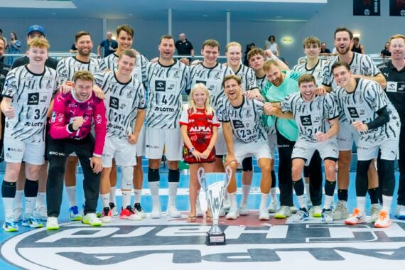 Eine Herren-Handballmannschaft posiert lächelnd mit einem Pokal auf dem Mittelkreis, zusammen mit einem kleinen Mädchen in einem roten T-Shirt. Die Gruppe wirkt glücklich und feierlich in einer Sporthalle, im Hintergrund sind Fans zu sehen.