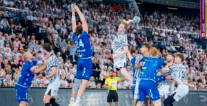 Ein Handballspieler in einem weiß-grauen Trikot springt zum Wurf hoch, während ein Spieler der blauen Mannschaft versucht, den Wurf zu blocken. Andere Spieler und eine große Zuschauermenge verfolgen das spannende Spielgeschehen in einer vollbesetzten Halle.